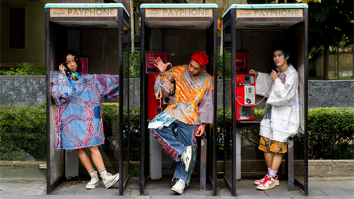 Three individuals in bold, colourful outfits pose inside vintage payphone booths set against a gritty, urban background.
