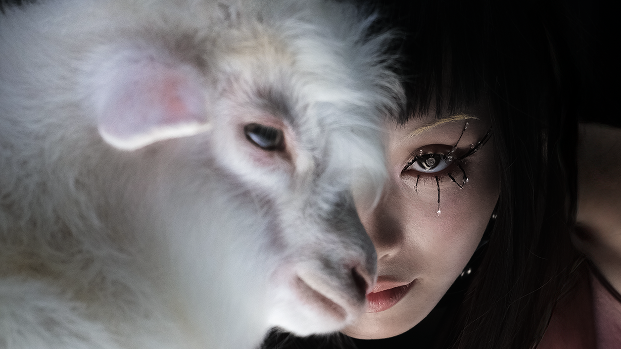 Close-up of a woman with crystal eye makeup beside a white goat, their faces aligned in soft lighting against a dark background.