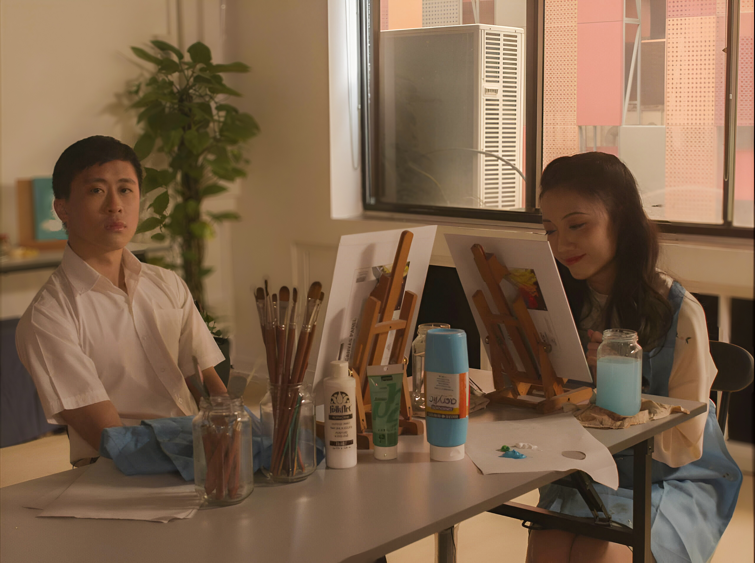 Two students paint on canvases in an art classroom, surrounded by brushes, paint tubes and water jars under soft natural light.