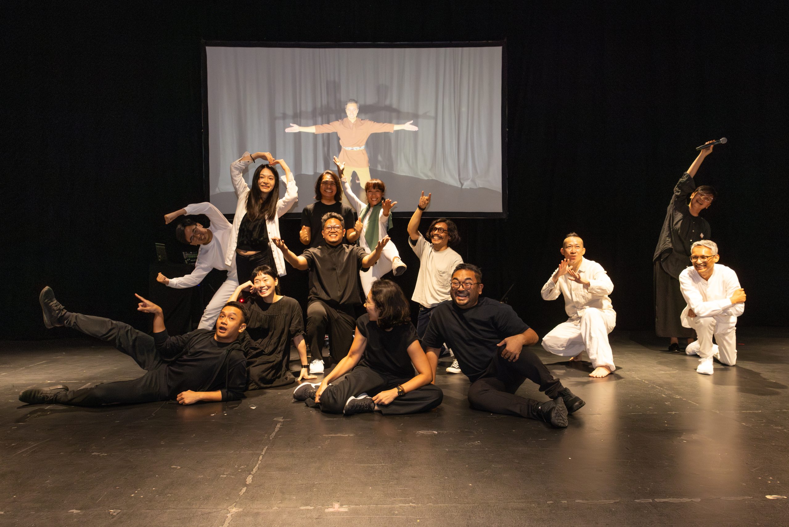 Group of performers strike playful poses on stage in front of a projection, celebrating the end of a performance with joyful energy.