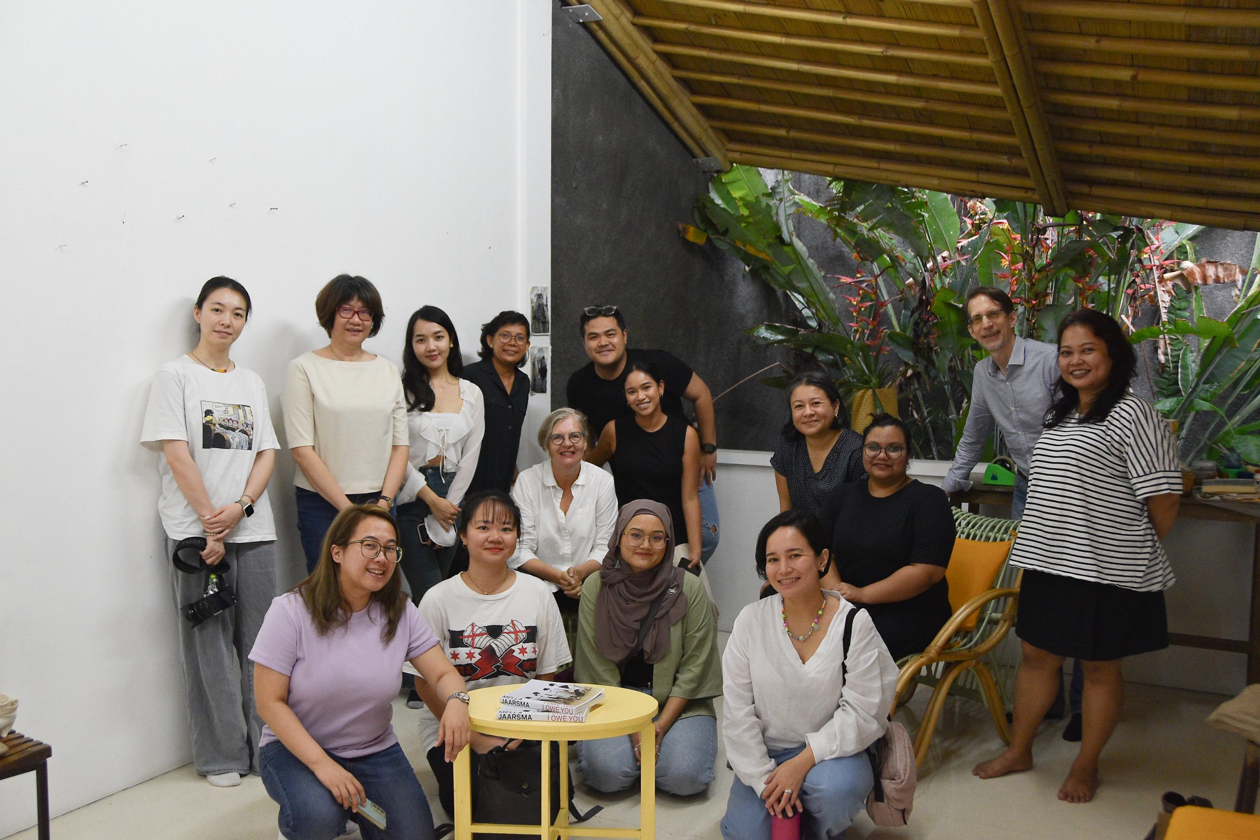 Fifteen people pose for a group photo in a cosy space with bamboo roofing, greenery and books during a community arts gathering.
