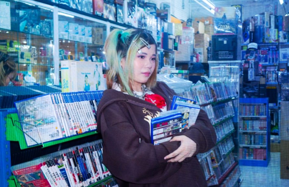 Young woman with dyed hair and clips stands in a brightly lit store holding game cases, surrounded by shelves of colourful media items.
