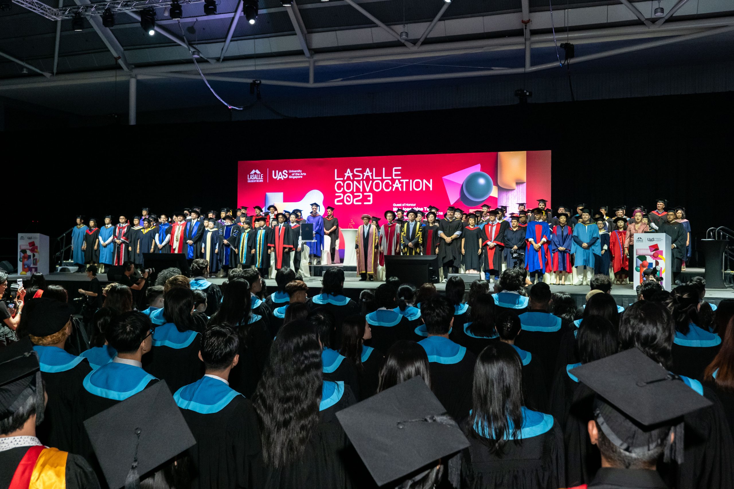 Graduates seated in front of a stage filled with robed speakers during LASALLE Convocation 2023, under bright stage lights.