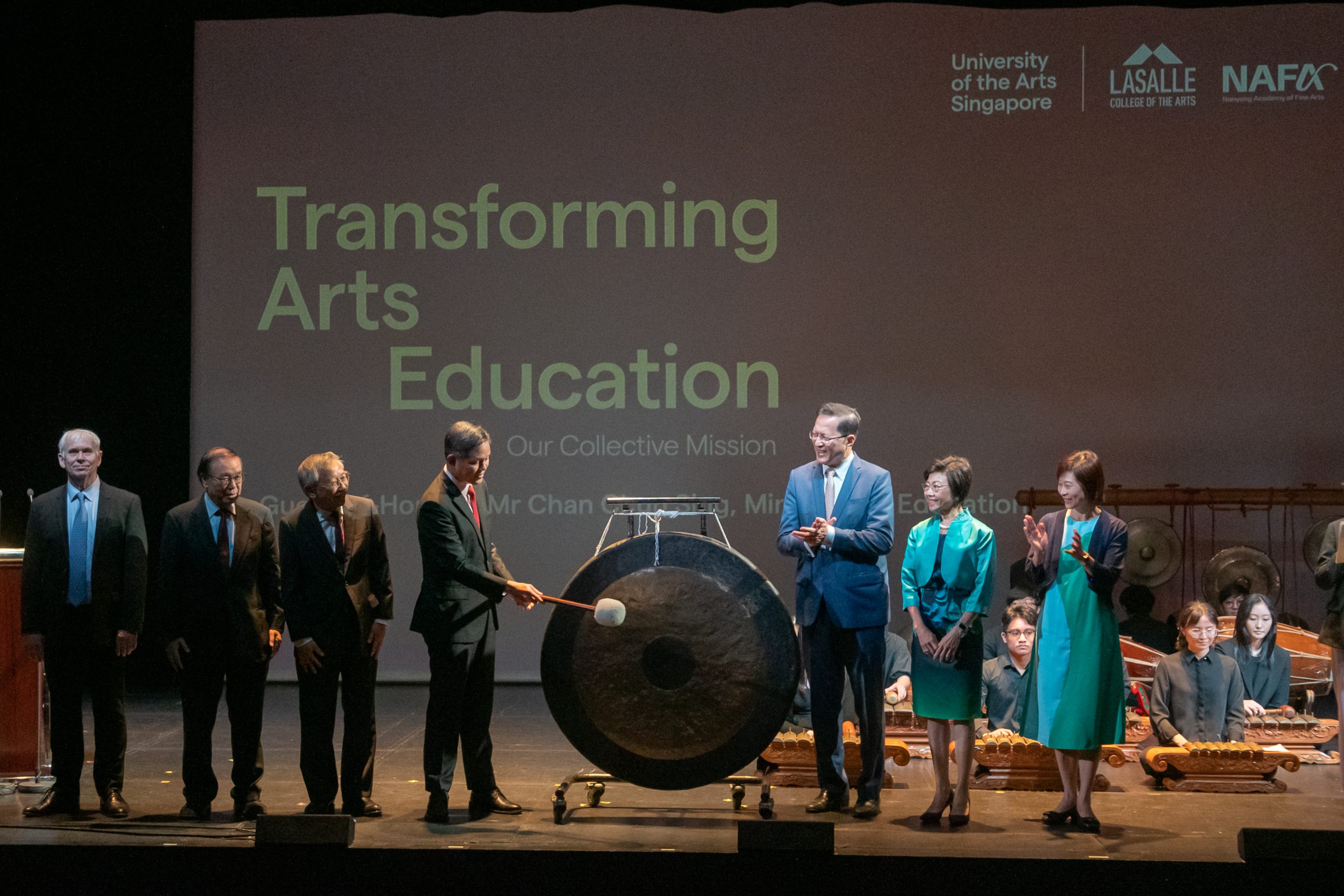 An official-looking man strikes a gong at an arts education event with UAS, LASALLE and NAFA leaders on stage, backed by a gamelan ensemble.