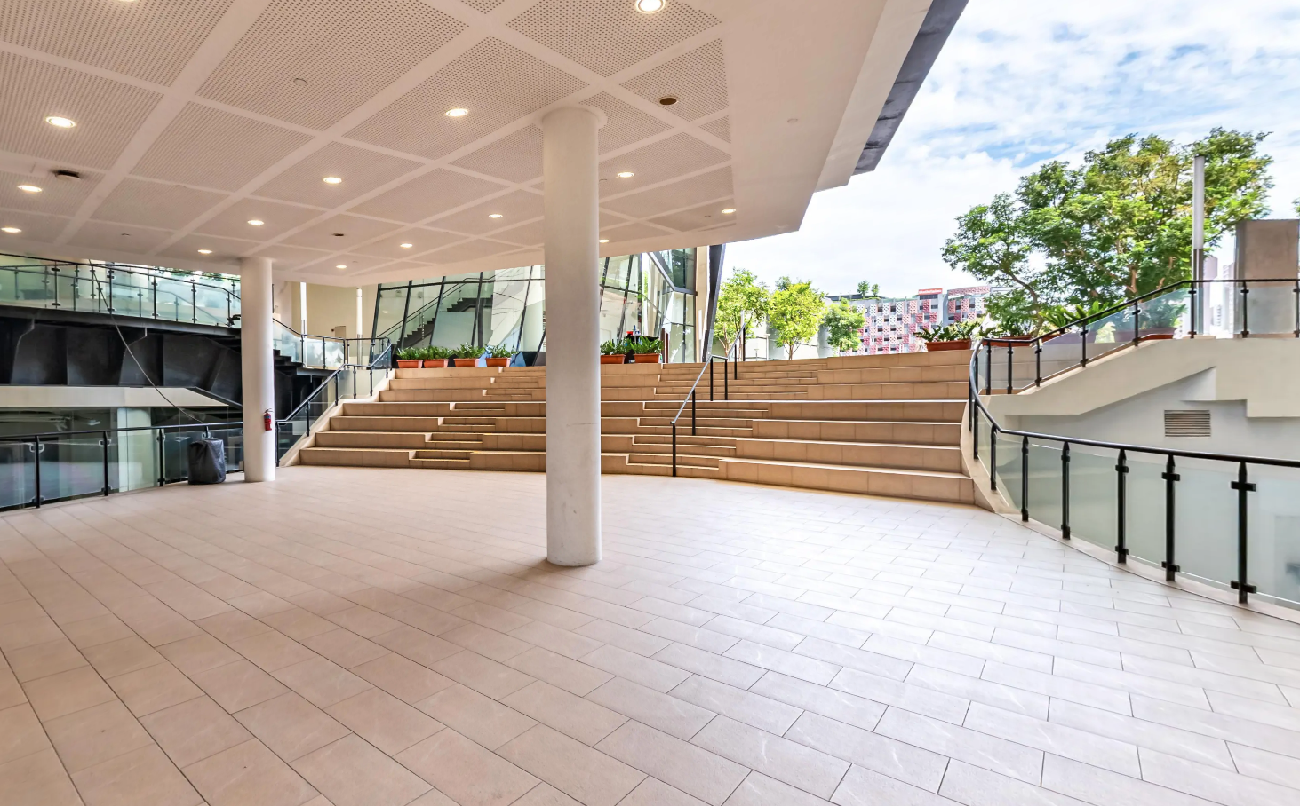 Tiered seating steps and open tiled plaza at LASALLE’s atrium, with glass balustrades and planted trees.