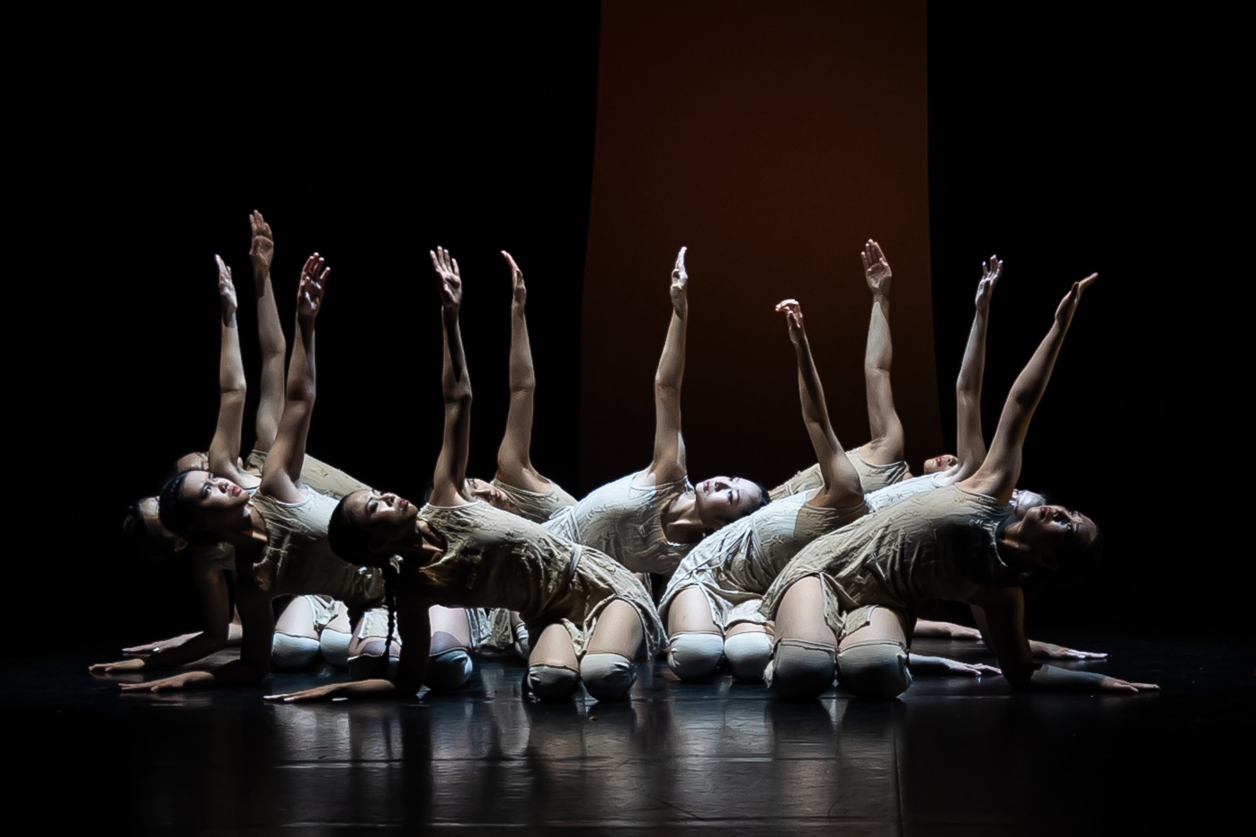 Group of dancers in beige costumes performing a synchronised movement on stage, with arms extended under theatrical lighting.