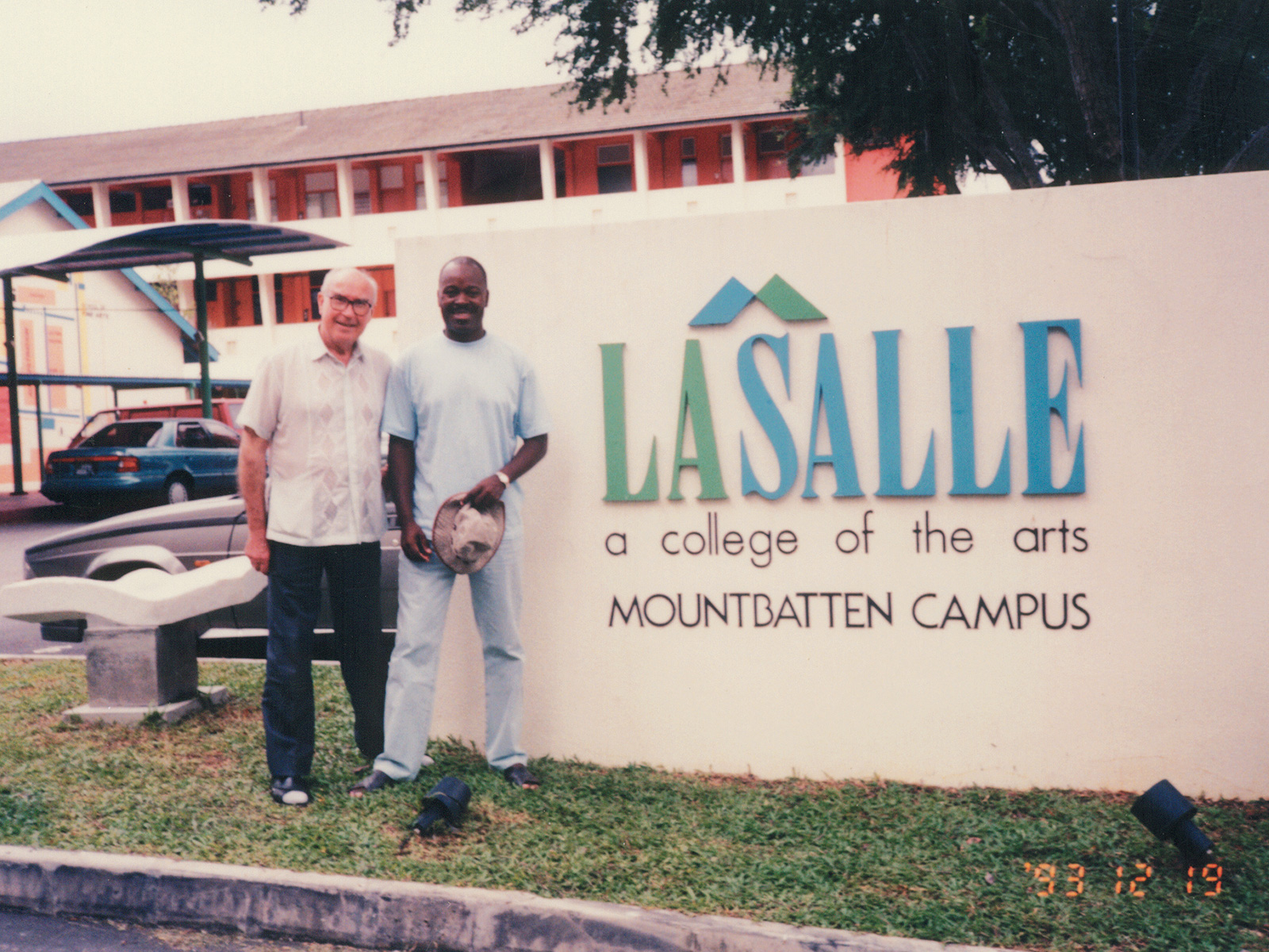 Vintage photography of two men pose beside the LASALLE Mountbatten Campus sign in the 1990s, with colourful school buildings and parked cars in the background.