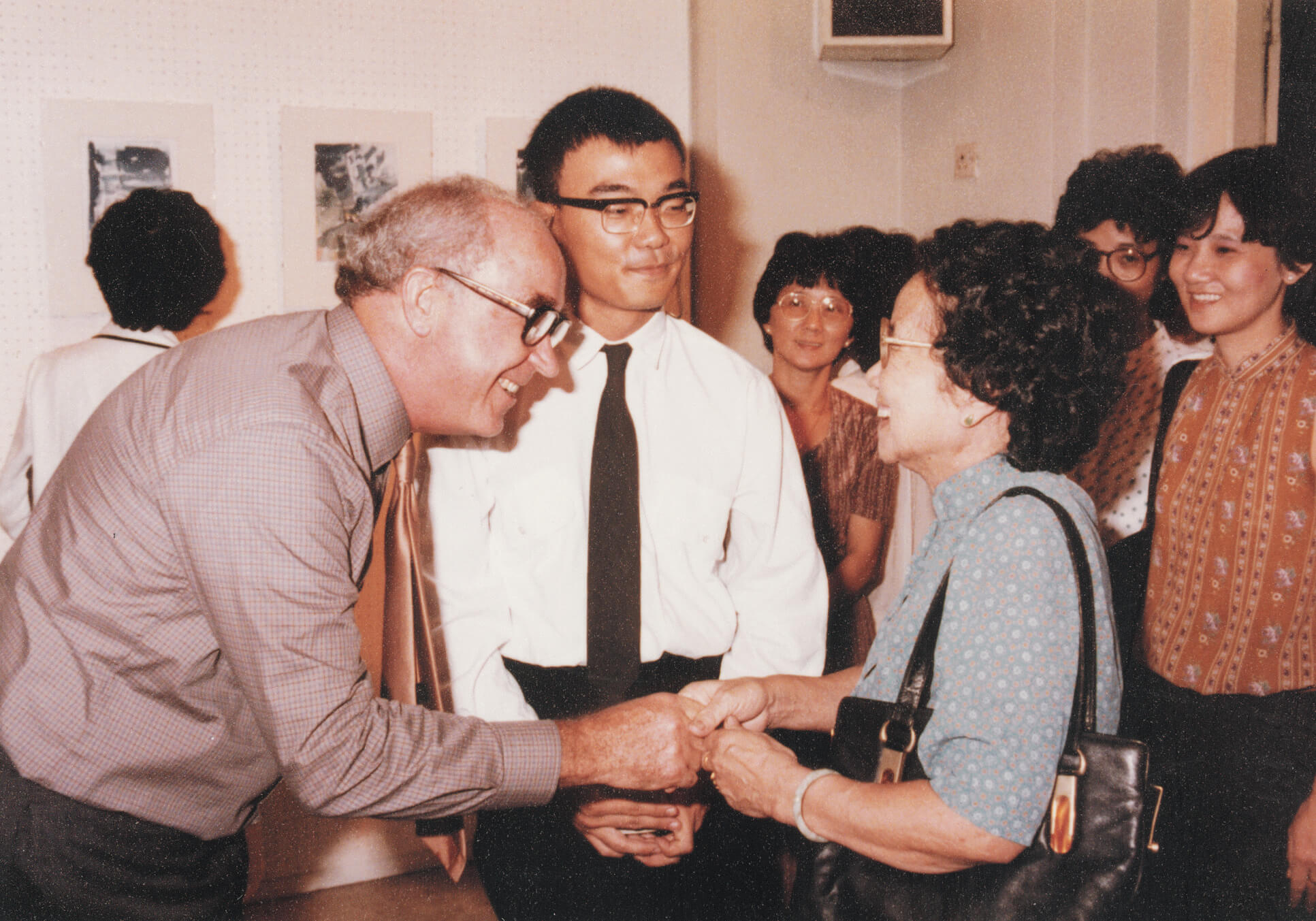 Vintage footage of Brother Joseph McNally warmly greeting a woman at an exhibition, surrounded by smiling guests in a gallery setting.