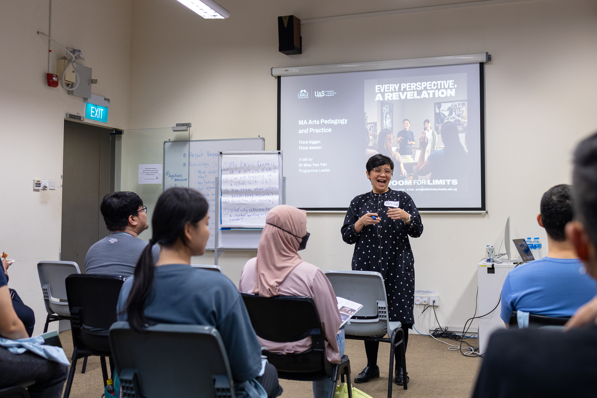 Lecturer speaks to diverse adult learners in a LASALLE classroom, with a projected slide, flip chart, and engaged students taking notes.