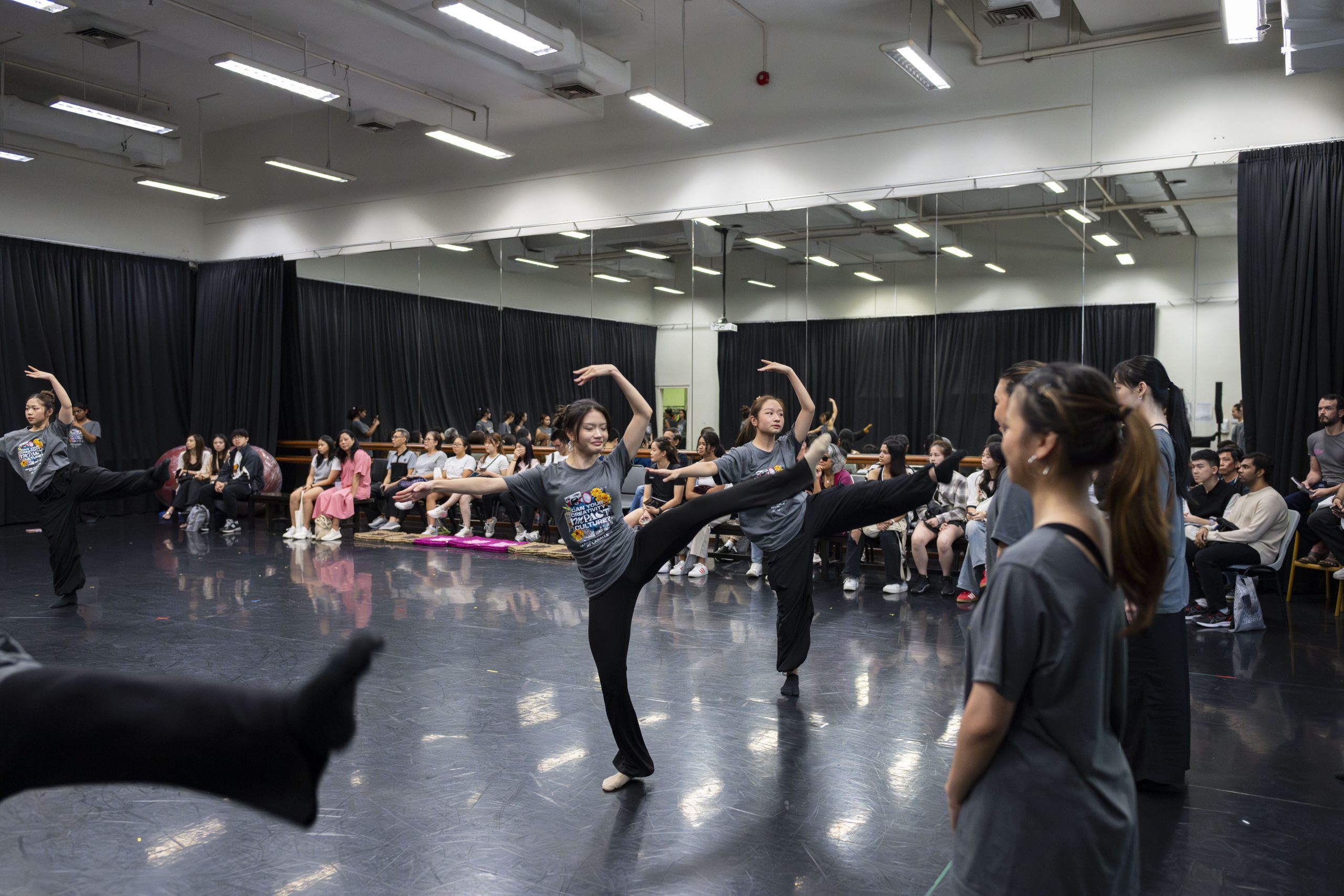 Dance students perform expressive movements in a mirrored studio as an audience watches from the side.