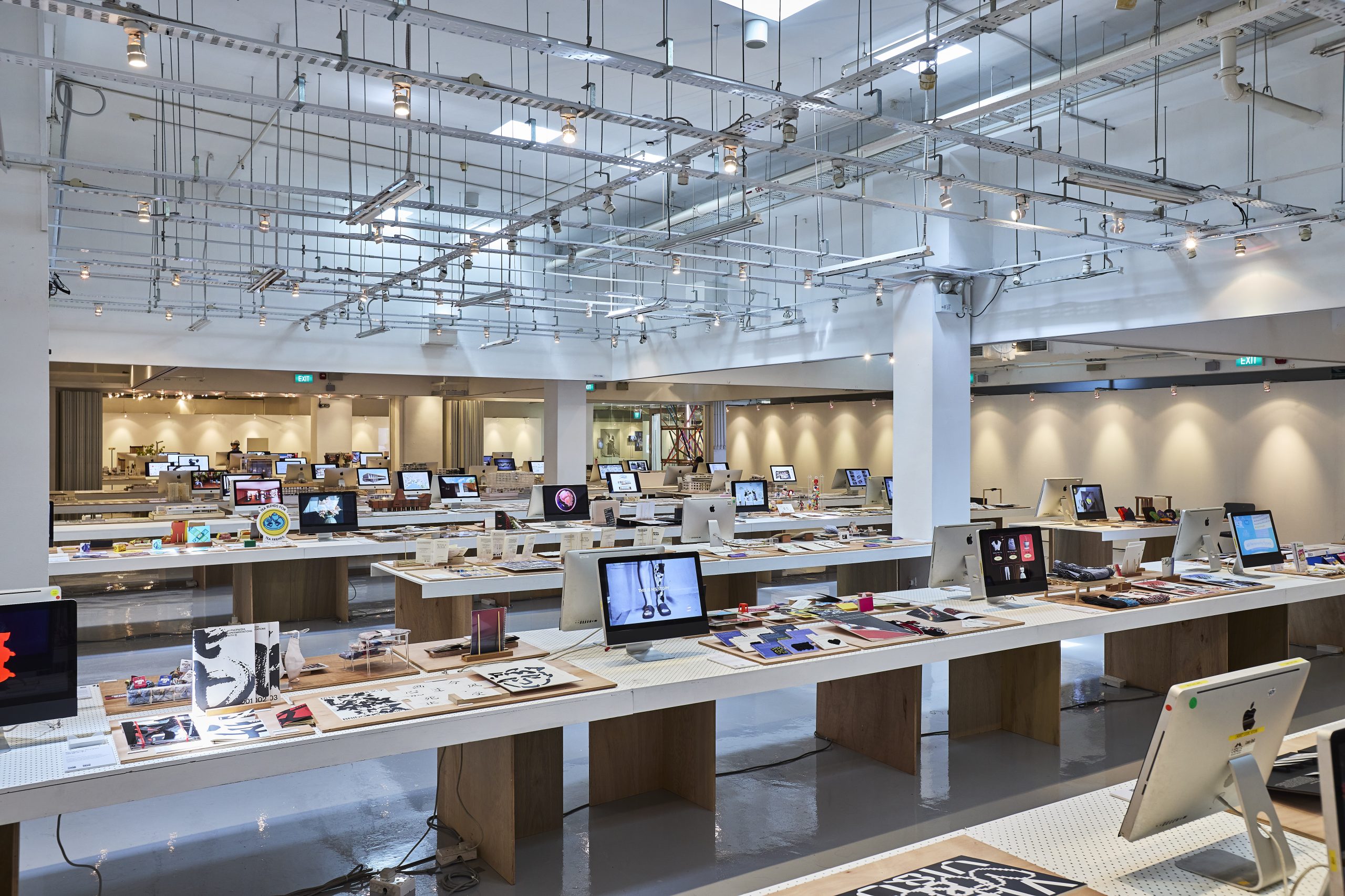 Showcase of student digital and design works displayed on long tables in a well-lit gallery space with desktop screens and printed portfolios.