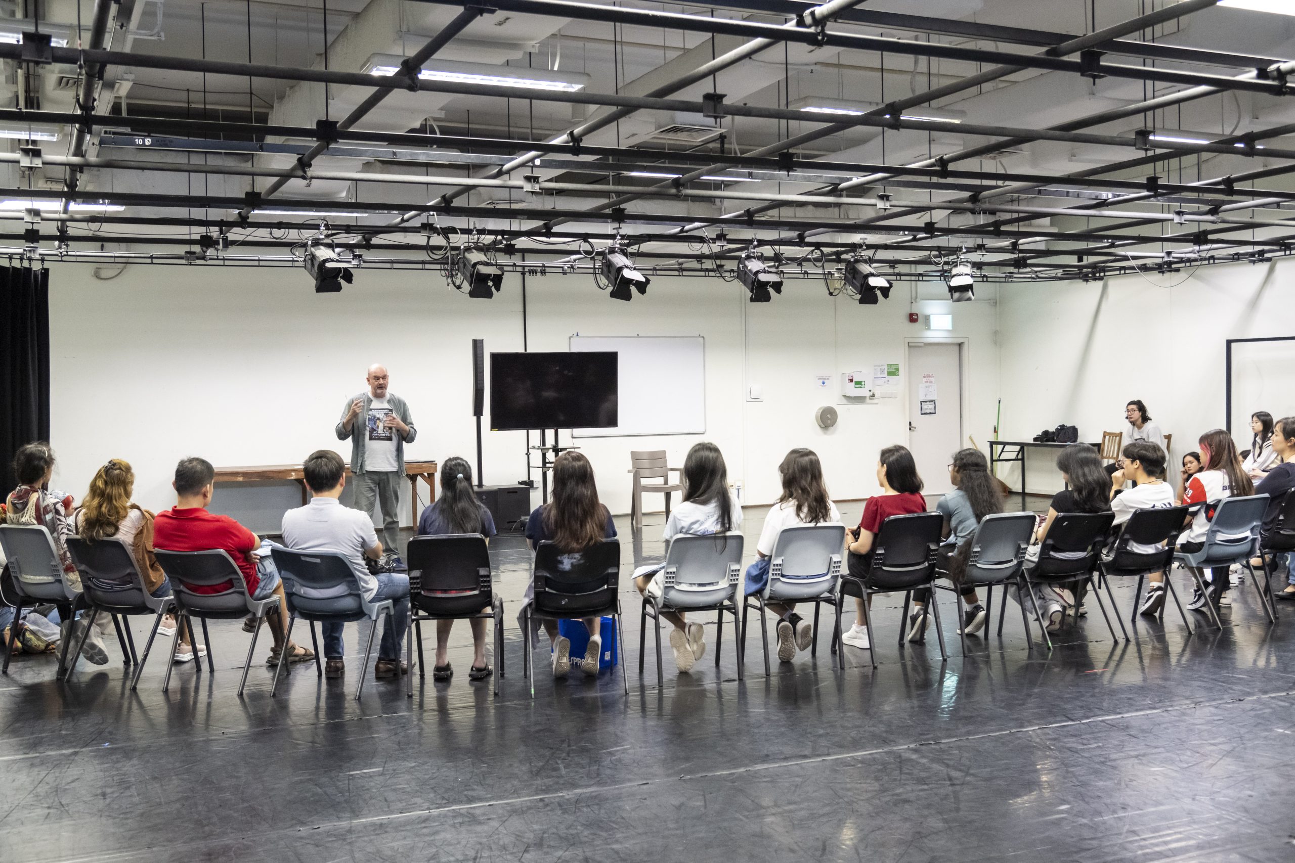 Lecturer speaking to a seated group of students in a black box theatre studio, with stage lighting rigs and a screen behind him.