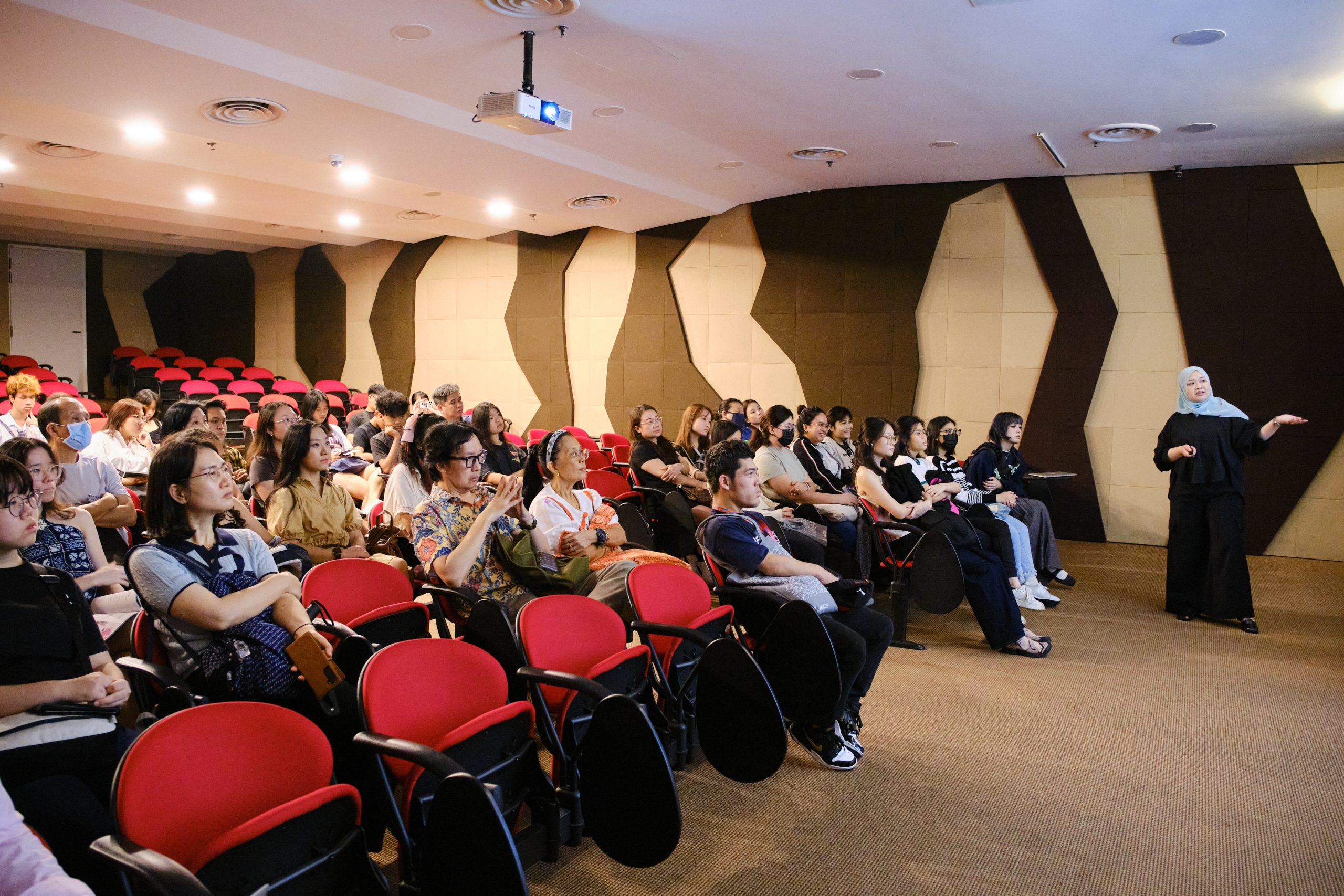 A speaker presents to a full auditorium of students seated in red chairs, inside an acoustically treated lecture room.