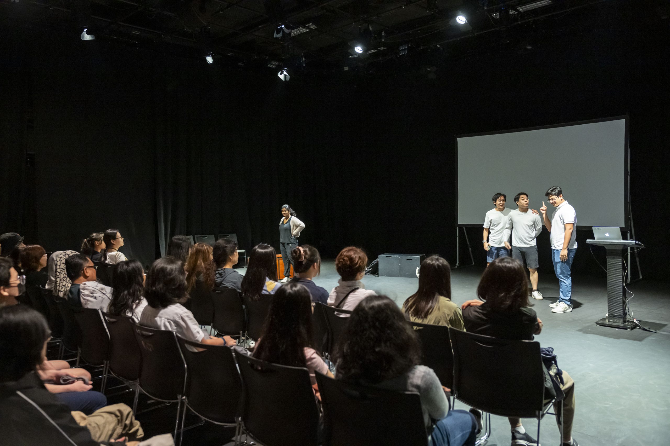 Students performing a short scene in front of an audience in a black box theatre, with a projection screen and podium on stage.