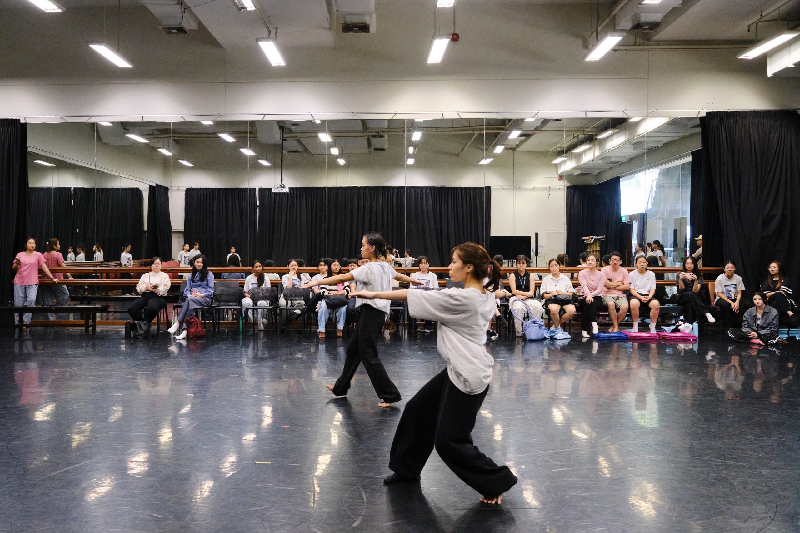 Two barefoot dancers in loose clothing perform in a large studio with mirrored walls, polished floor, and seated audience.