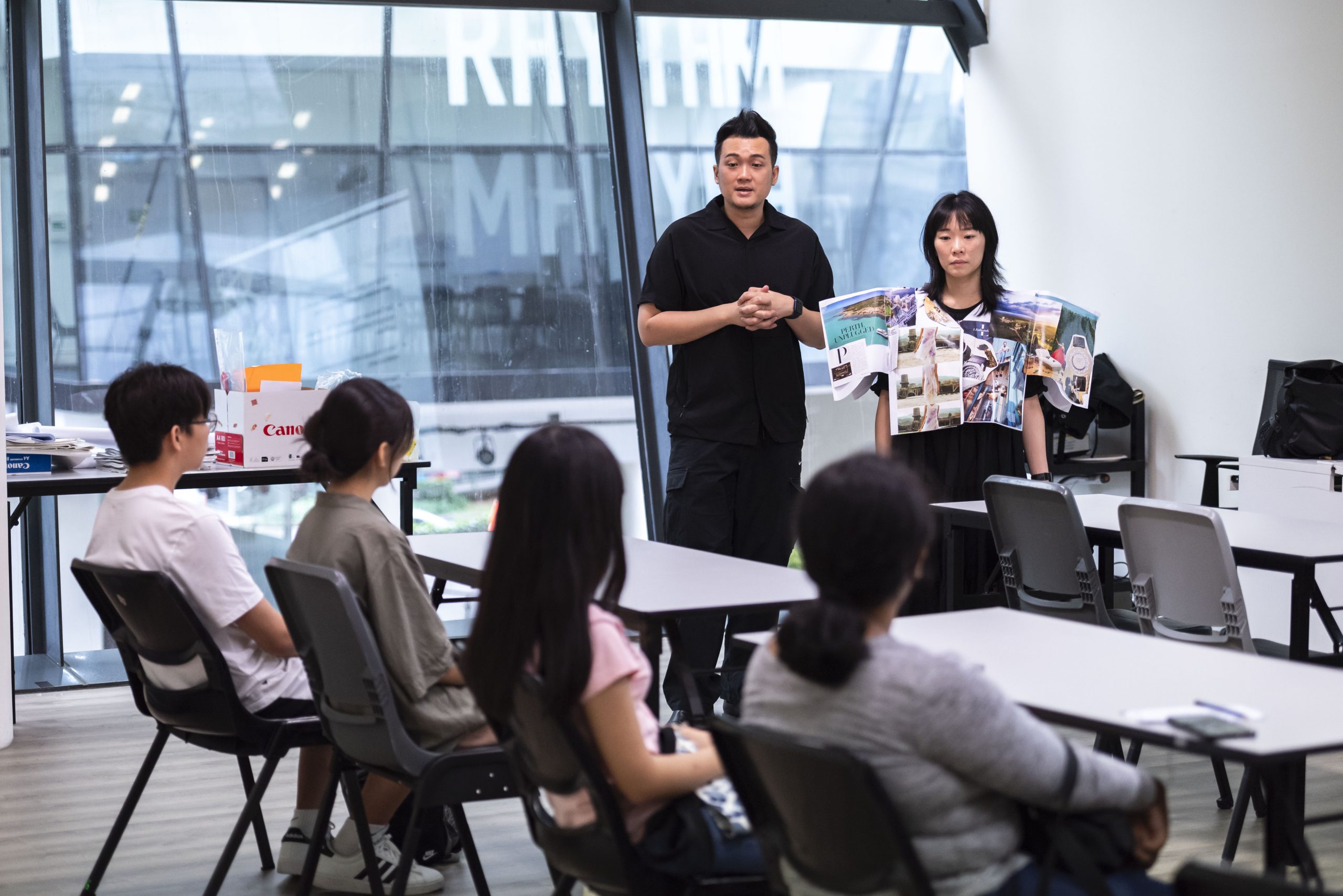 Two LASALLE students presenting a creative wearable seemingly made from brochures to a seated classroom audience in a design studio.