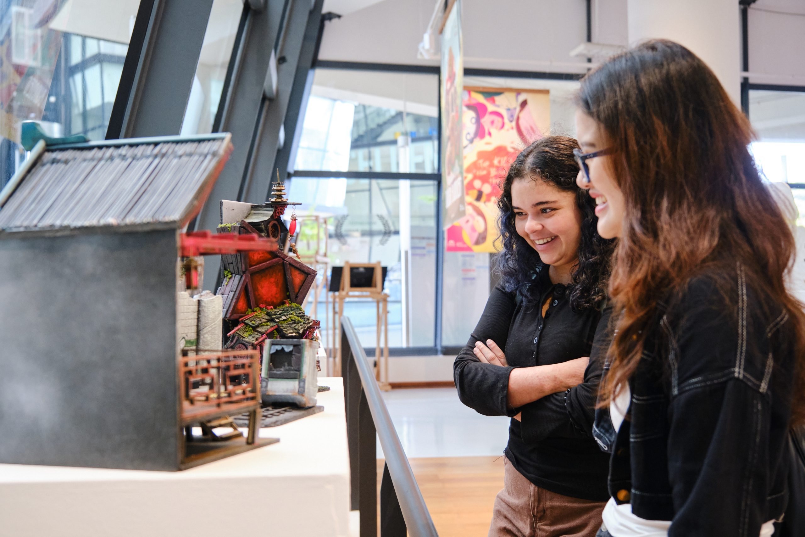 Two LASALLE students smiling while admiring a detailed architectural model of a fantasy-style house on display inside a brightly lit campus gallery.