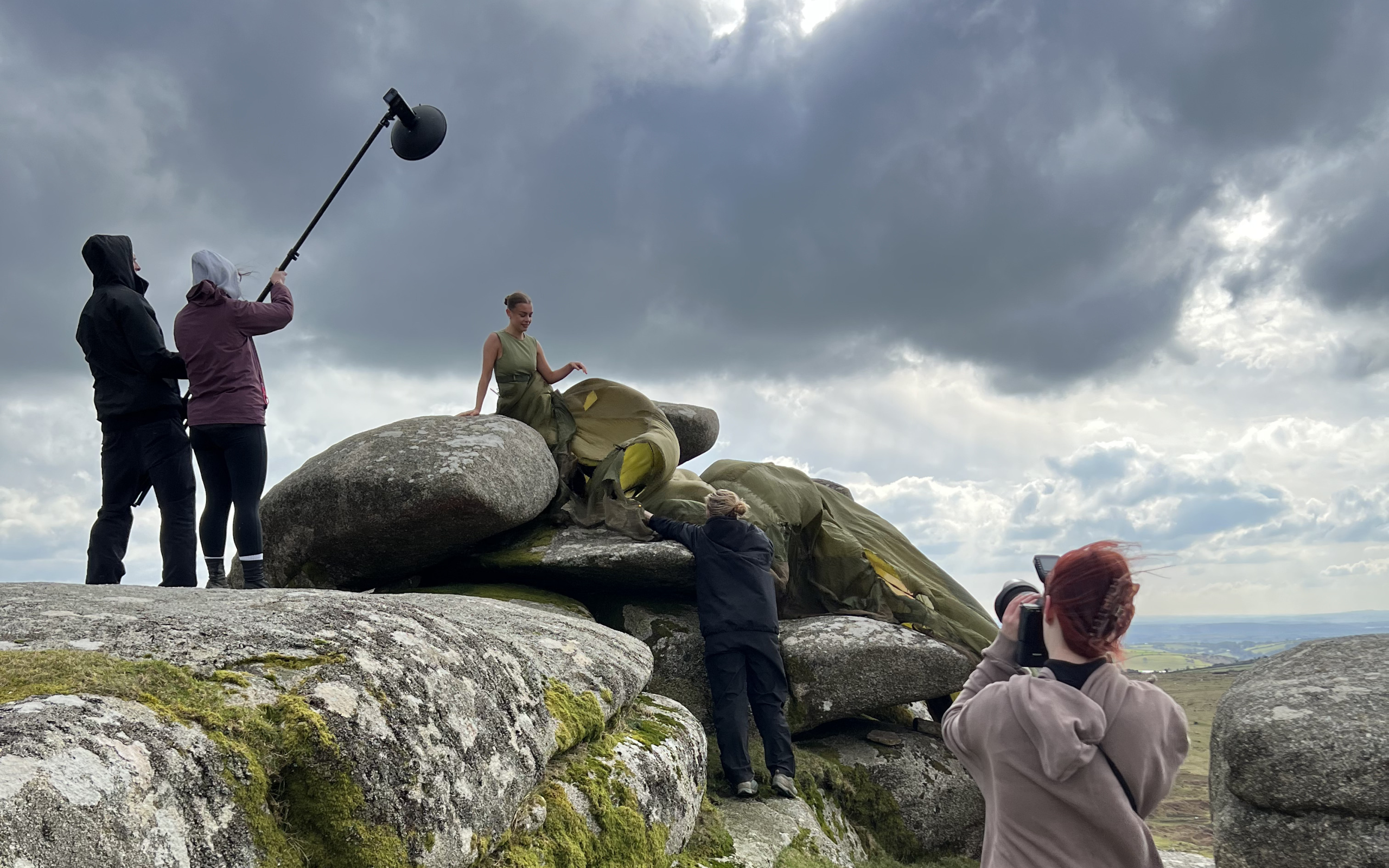 Behind-the-scenes of a fashion shoot on mossy rocks under dramatic clouds, with a model in a flowing green dress and crew adjusting lighting and capturing photos.