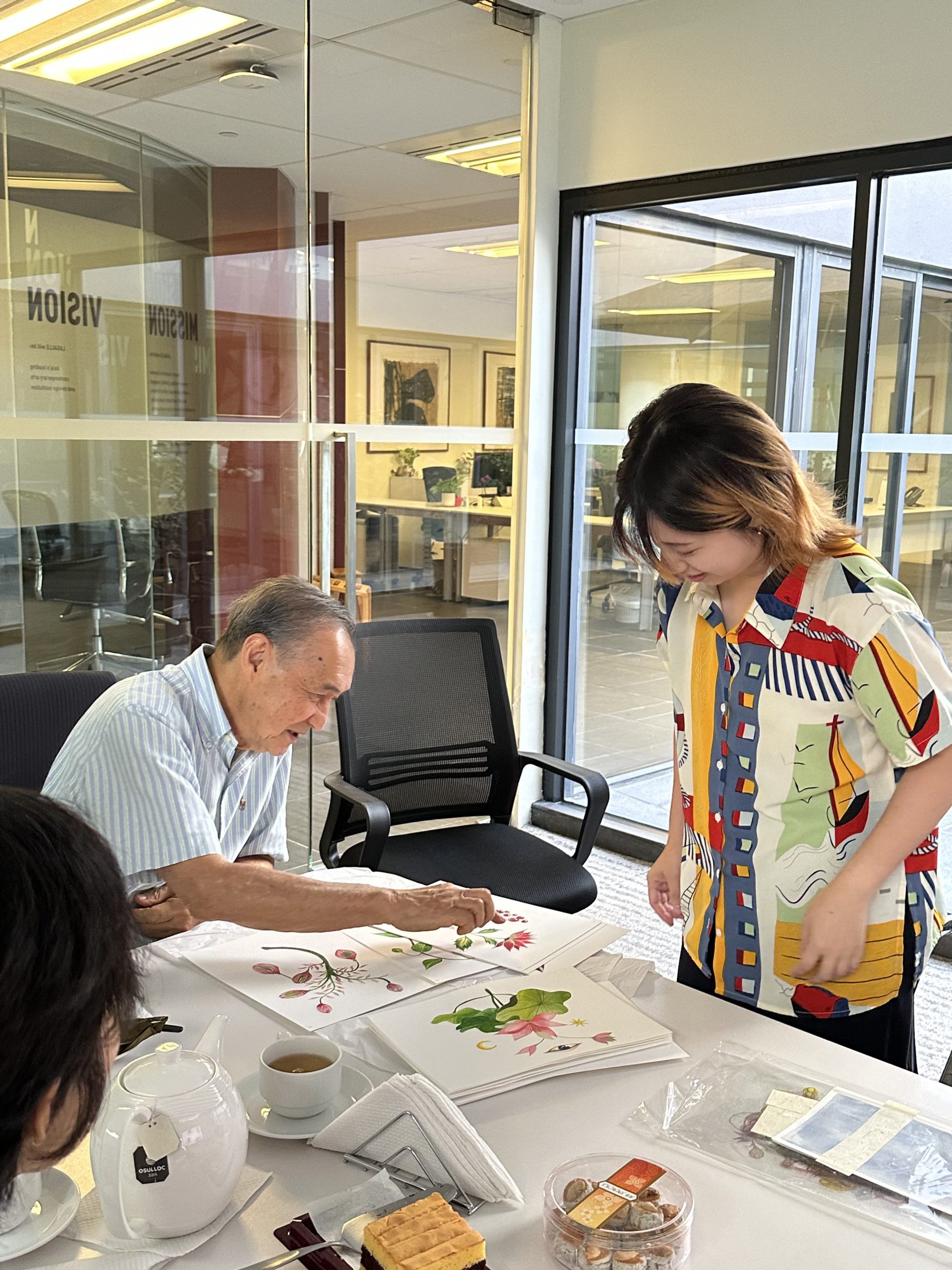 An elderly man demonstrates Chinese brush painting of flowers while a younger woman observes, in a room with tea and snacks on the table.
