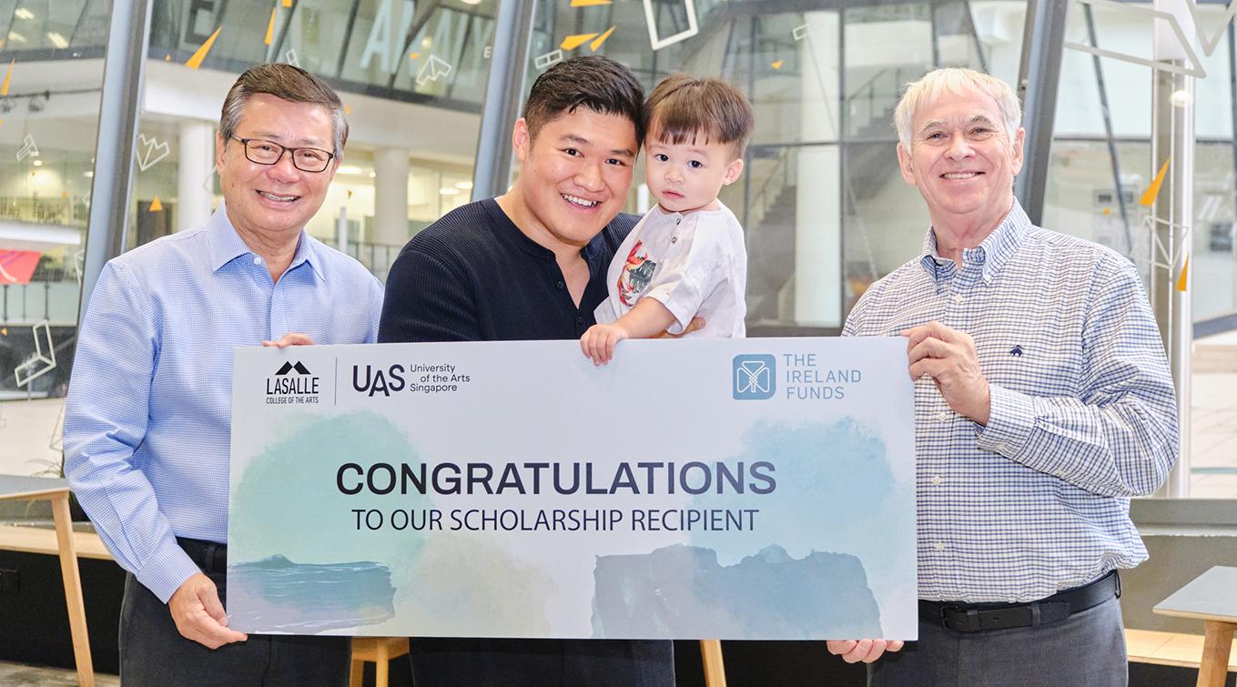 Scholarship recipient holding a congratulatory banner with his child, flanked by LASALLE and UAS representatives at an award ceremony supported by The Ireland Funds.