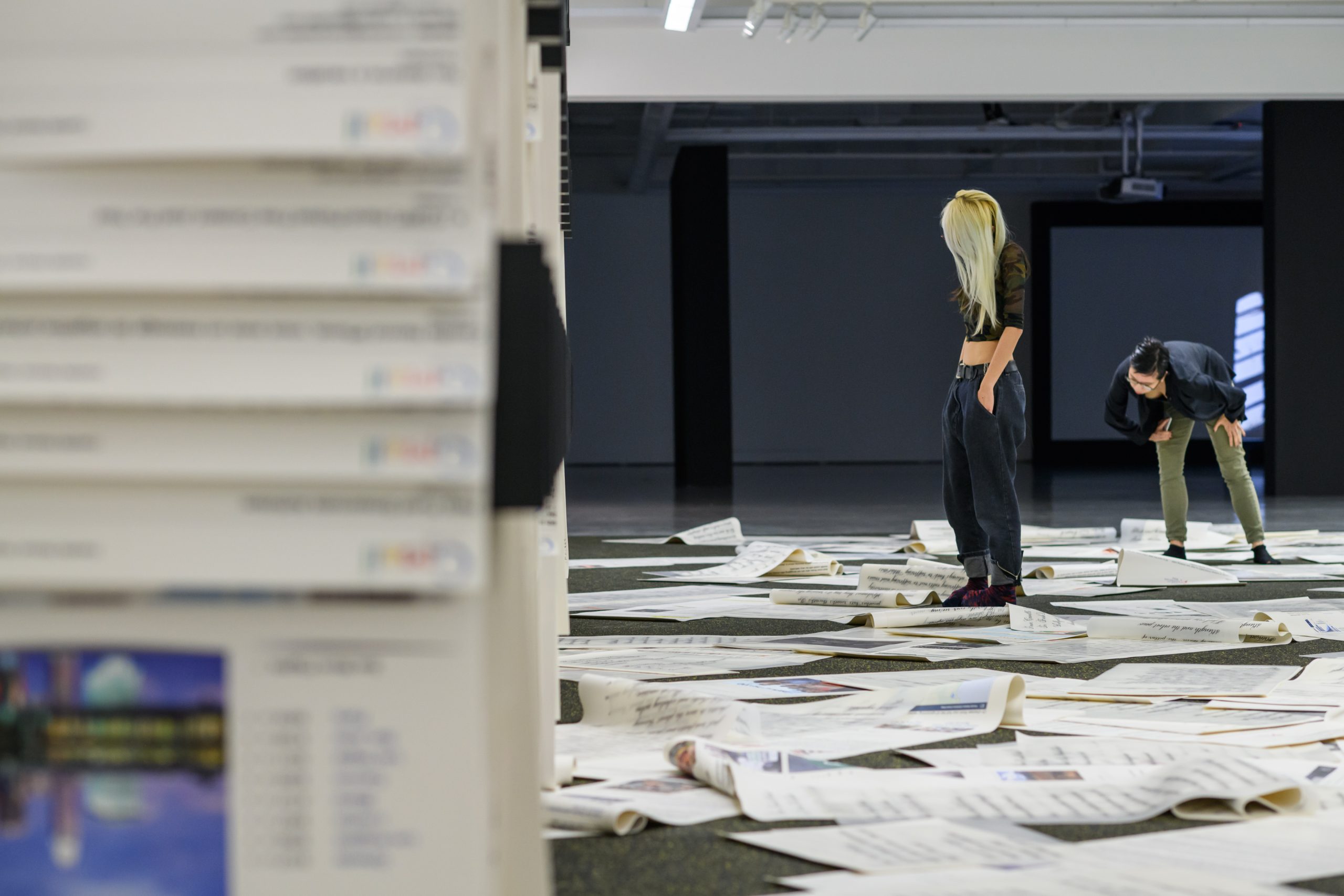 Two visitors walk among large sheets of printed text and images spread across the LASALLE gallery floor in an installation.