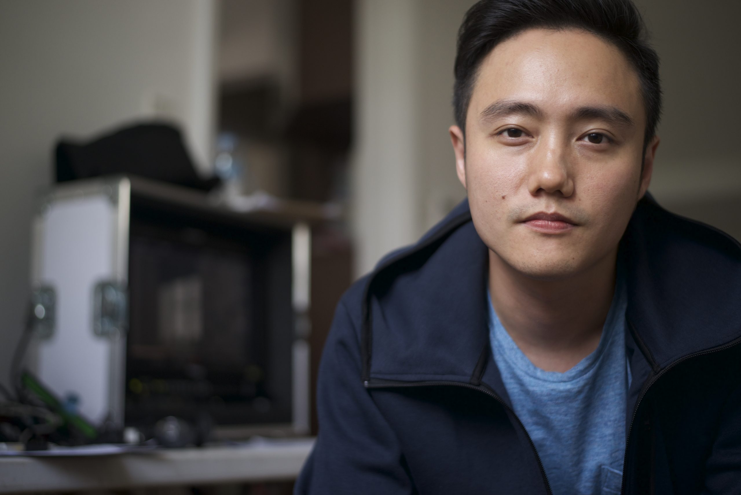 Portrait of a young man in a blue shirt and jacket, seated indoors with blurred equipment and shelves in the background.