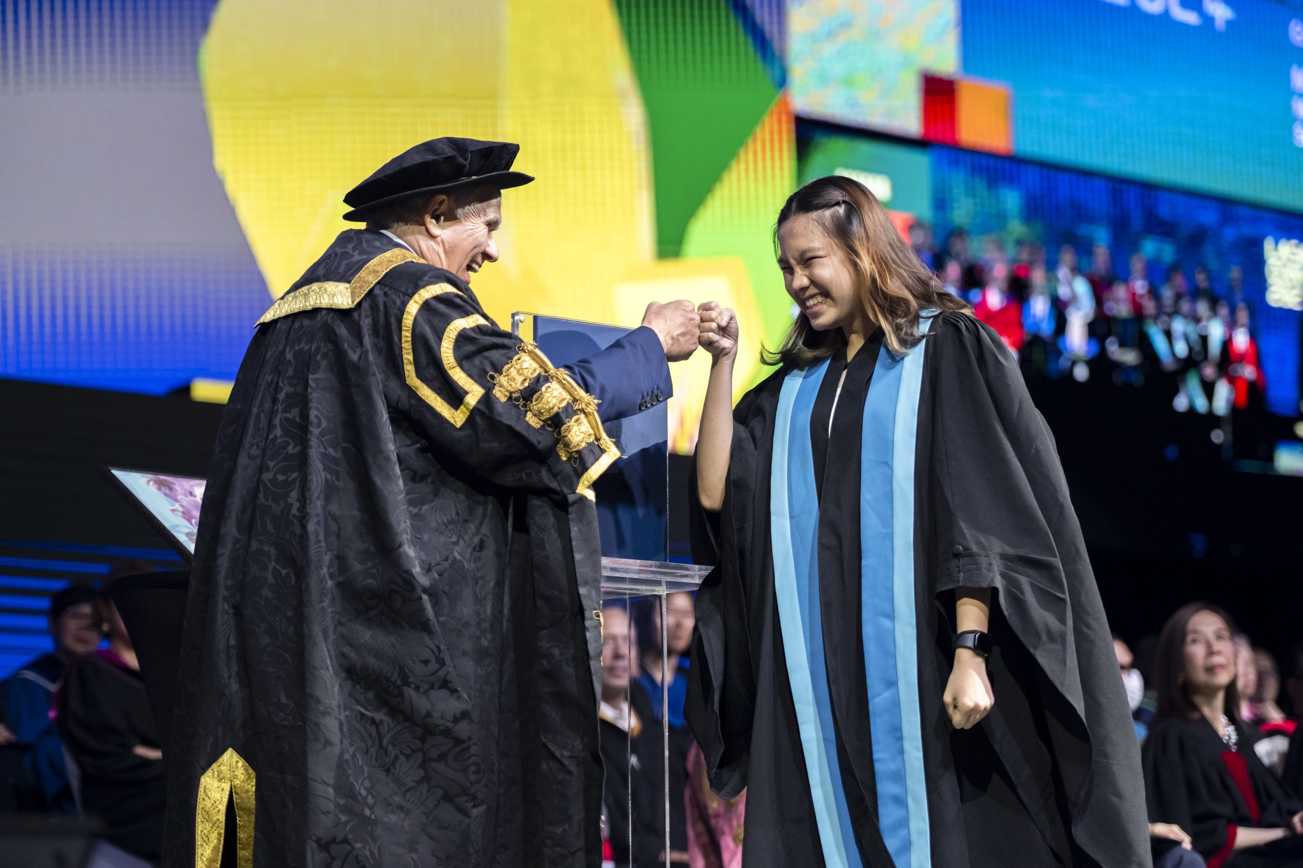 Smiling graduate in gown and sash fist-bumps a LASALLE's President onstage at a graduation ceremony.