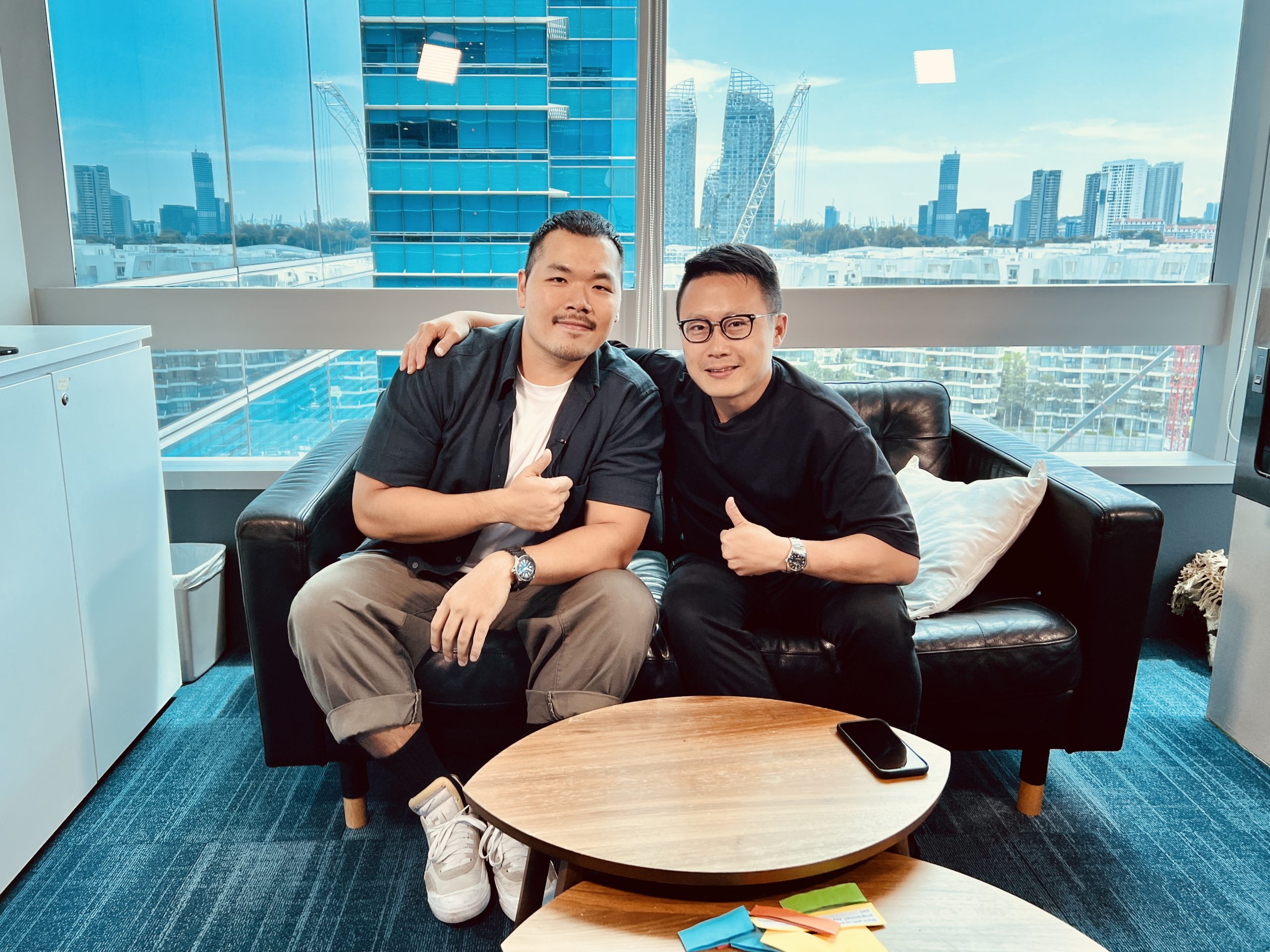Two men sit on a black leather sofa in an office with city views, smiling and giving thumbs up beside a round table with a phone and notes.