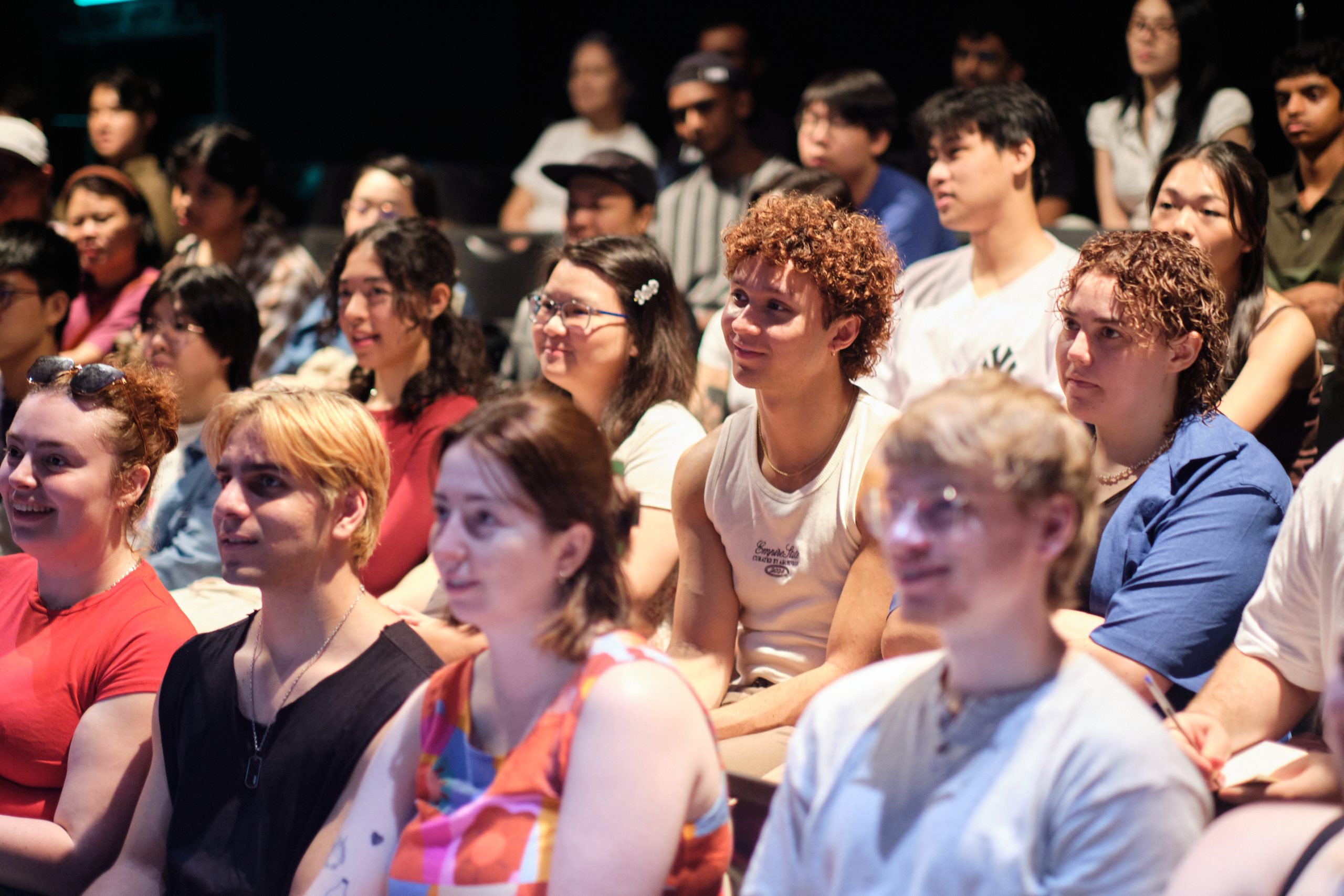A diverse group of students sit closely together in an auditorium, smiling and focused on a presentation.