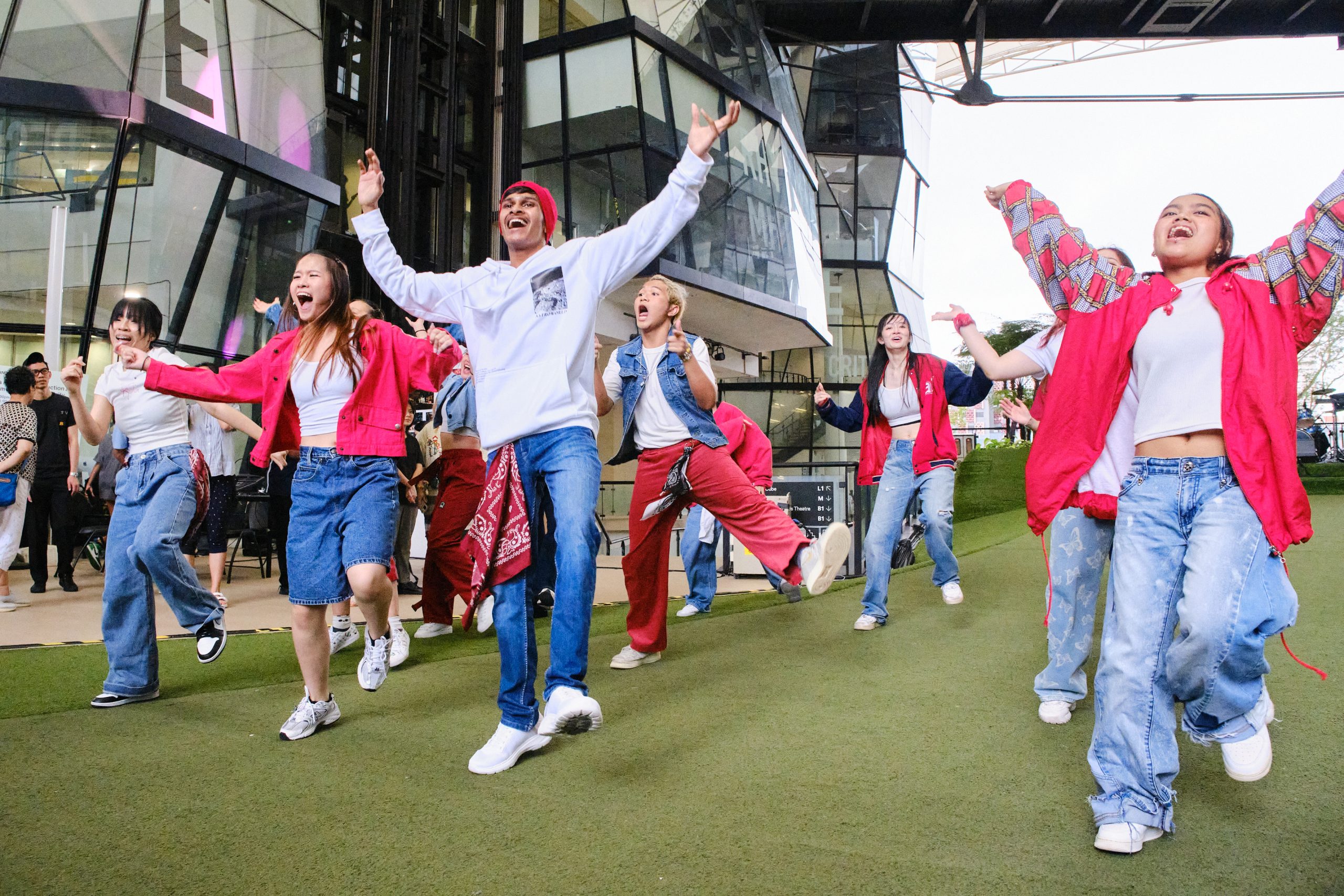 A group of young performers in red and white outfits dancing energetically on green turf outside a modern glass campus building.
