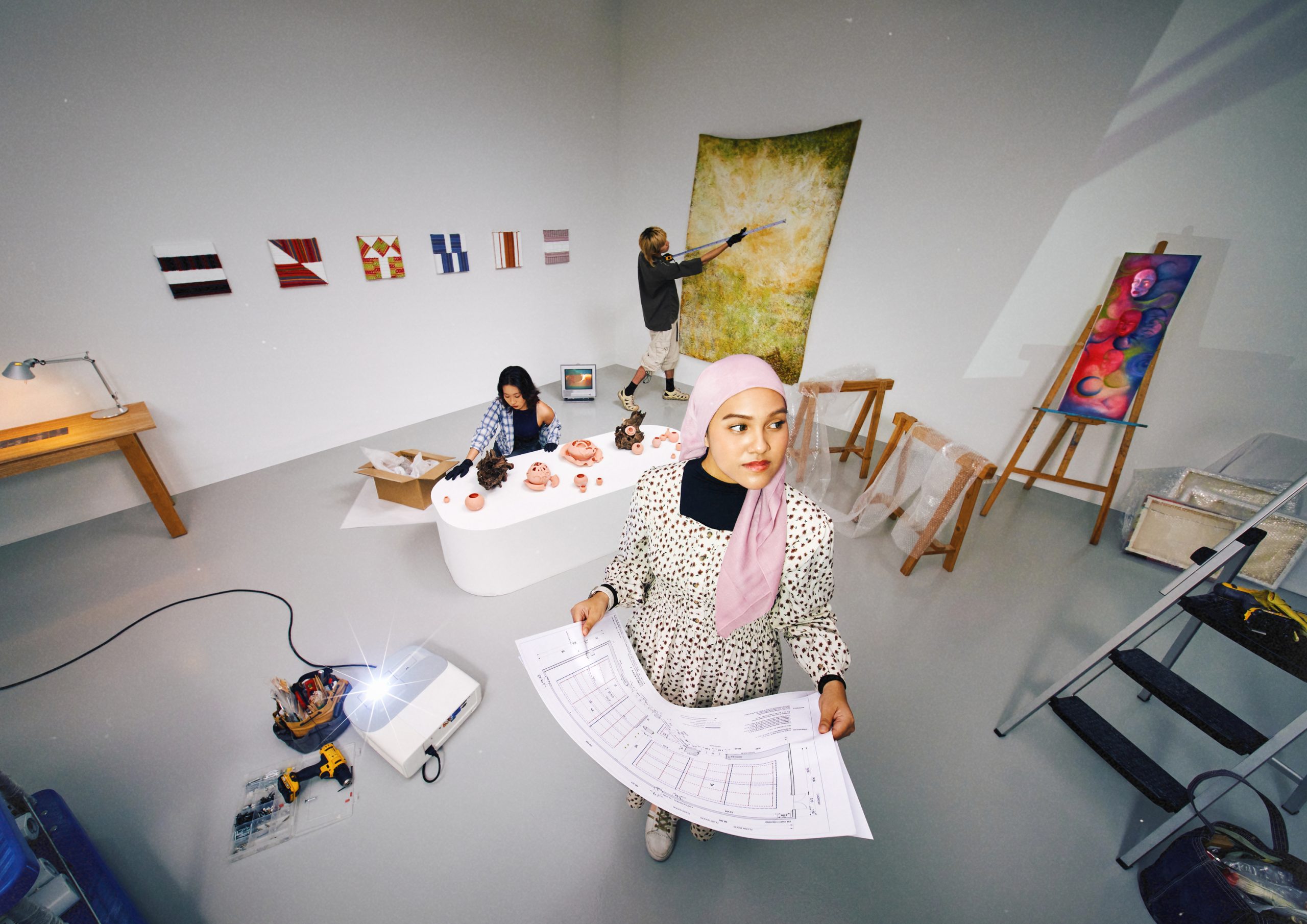 Three students setting up an exhibition space with ceramics, paintings and tools, while one foreground figure in a pink headscarf holds architectural floor plans.