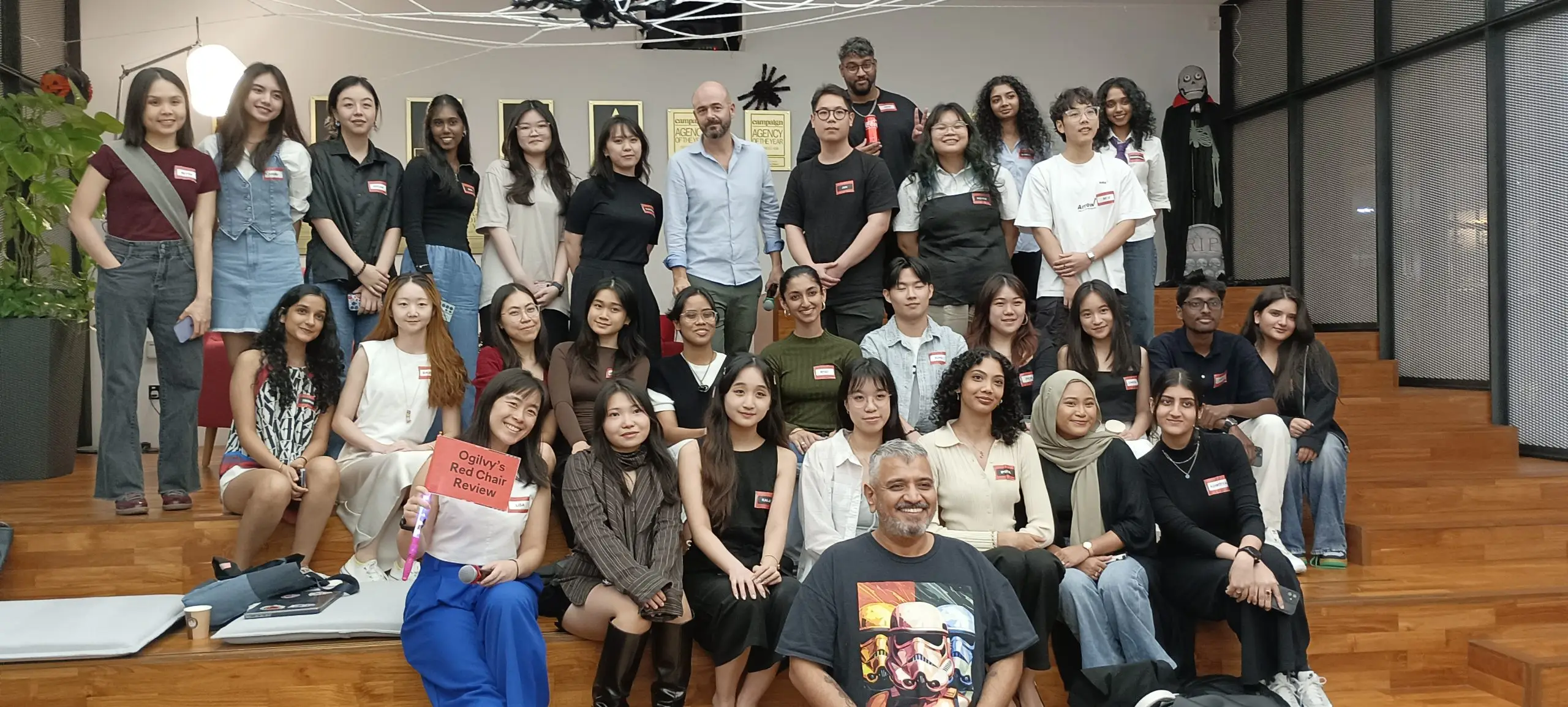 Group photo of students and professionals seated on tiered wooden steps at Ogilvy’s Red Chair Review, with Halloween décor in background.